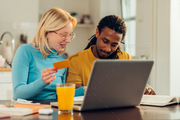 A happy multicultural couple is at their home and using a credit card for paying bills and taxes