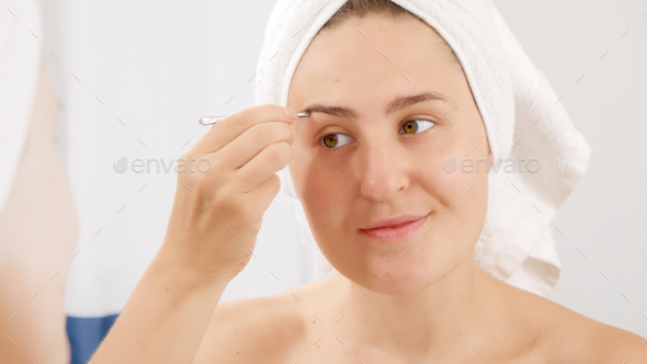 Young woman in bath towel using tweezers for plucking eyebrows