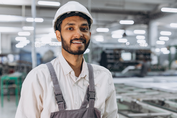 Industrial Indian international worker indoors in factory. Young technician in white hard hat.