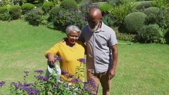 African american senior couple smiling while watering plants together in the garden alt