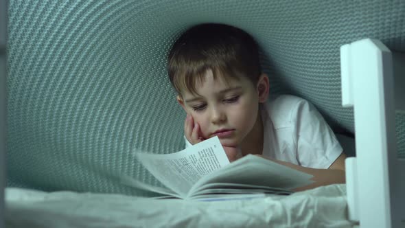Little, Cheerful, Seven Years Old Boy Lying on His Belly on the Sofa, with Open Book in Front of Him