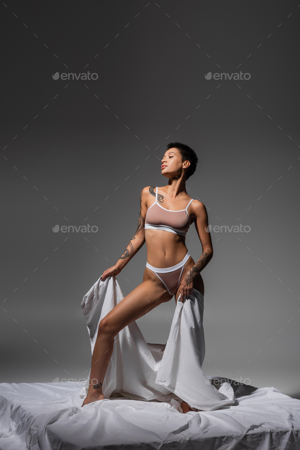 young and provocative tattooed woman with sexy body and short brunette hair  posing in beige lingerie Stock Photo by LightFieldStudios