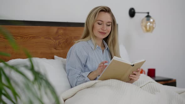 Young Charming Intelligent Caucasian Woman Reading Book Sitting in Bed Smiling alt