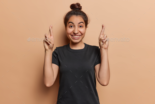 Photoo of cheerful Iranian girl keeping fingers crossed looks gladfully at camera dressed in casual