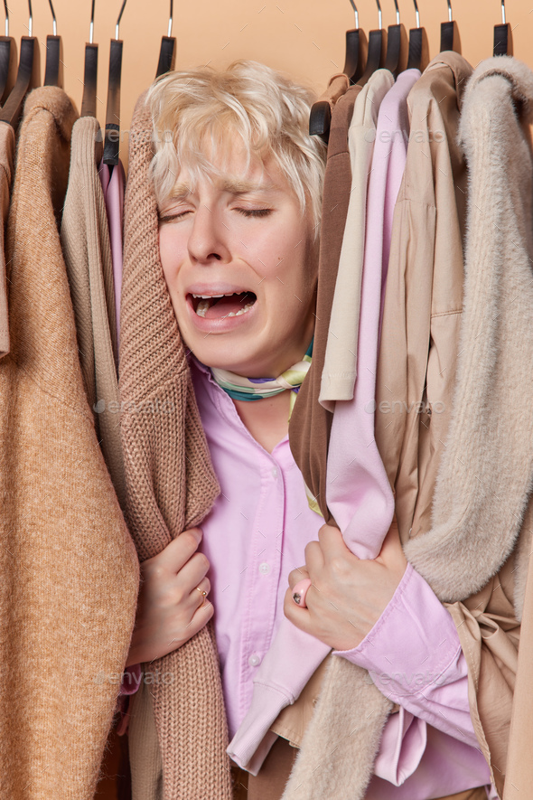 Doleful fair haired woman poses around clothes on hangers cries from despair keeps mouth opened