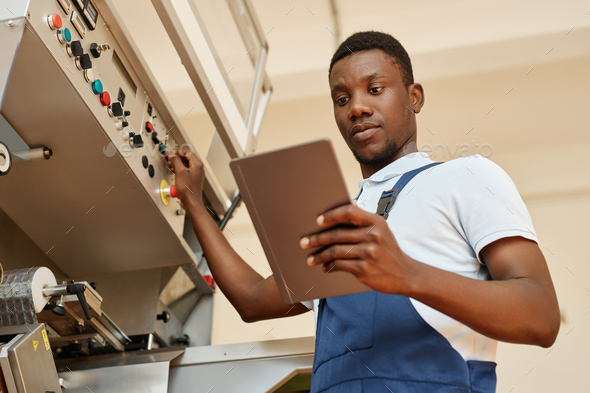 Black man as factory worker holding tablet operating machine unit