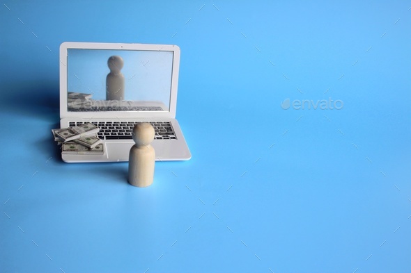 Wooden doll in front of laptop and pile of money. Blue background with copy space.