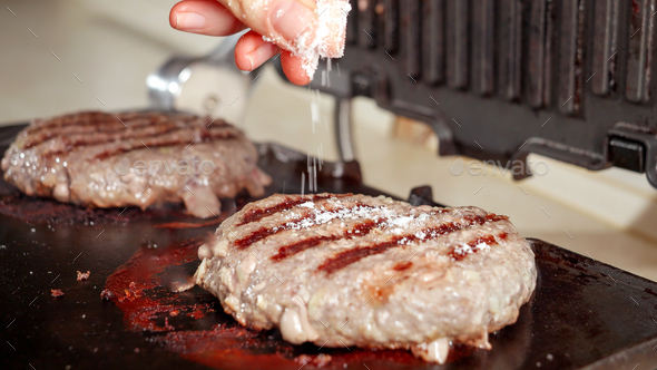 Closeup of hand salting beef burgers cooking on electric grill at