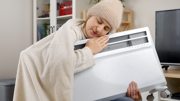 Young woman covering in blanket and holding electric heater to warm herself in cold apartment
