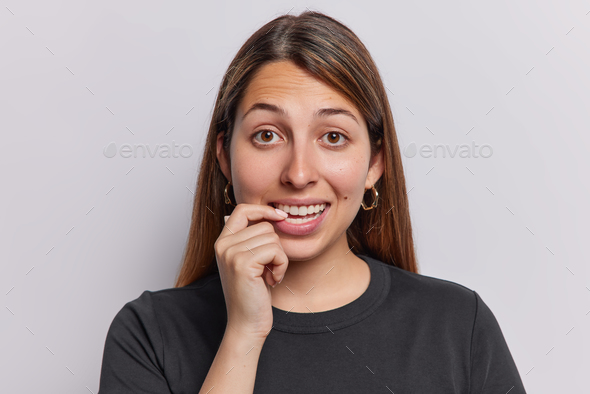 Portrait of curious long haired woman bites finger looks with intriguing expression wears casual