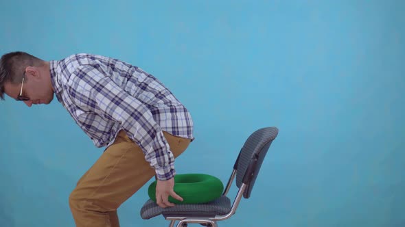 Positive Young Man with Glasses Sitting on an Orthopedic Pillow From Hemorrhoids on a Blue alt