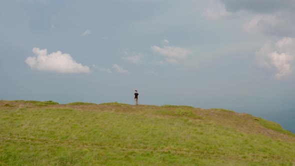 Aerial Drone View. Flying Around Young Man Standing on Top of the Mountain at Sunset alt