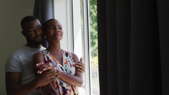 African american couple embracing each other while looking out of the window at home alt