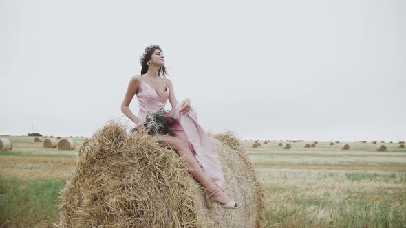 Carefree Lady in Dress Posing for Camera on a Haystack in Windy Field alt