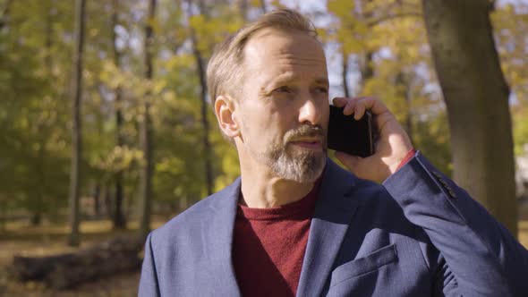 A Middleaged Handsome Caucasian Man Talks on a Smartphone with a Smile in a Park in Fall