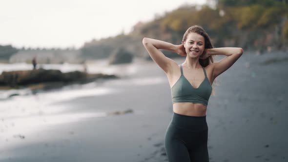 Woman in Sportswear Walks Along Black Sand Beach Looks at Camera and Smiles alt
