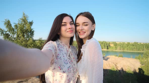 Two Pretty Young Passionate Women in Dresses in Mountains with Blue Lake in Background Make Selfie alt