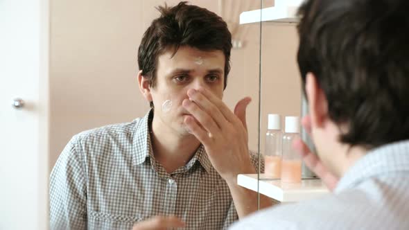 Man Puts Cream on His Face After Shaving While Looking in the Mirror. Brunette in a Plaid Light