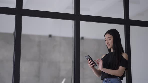 Positive Confident Asian Woman Messaging Online in Smartphone App Standing at Glass in Office in alt