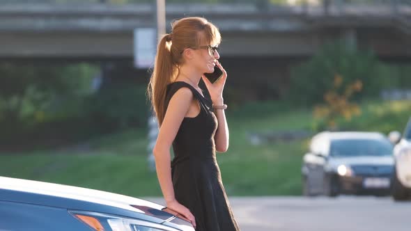 Young Female Driver Standing Near Her Car Talking on Mobile Phone on a City Street in Summer alt