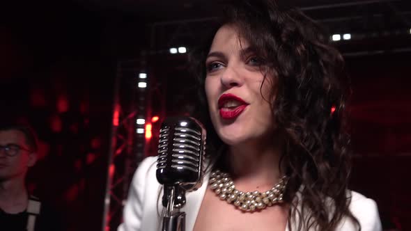 A Stylish Girl Vocalist Sings on Stage in a Vintage Microphone. In the Background Musicians Play alt