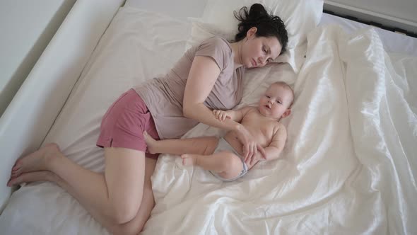 Happy Young Mother Lying with Cute Infant Toddler Baby Boy on Bed Holding Him on Arms Hugging and