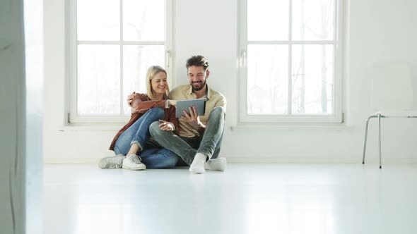 A couple sitting on a white floor in a loft looking in a tablet
