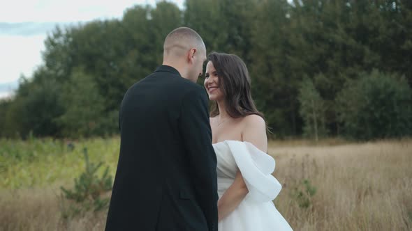 Happy Bride and Groom Hold Hands and Look Into Each Other's Eyes Against the Backdrop of a Field and alt