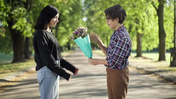 Side View Loving Teenage Nerd Boy Talking Giving Bouquet of Flowers to Girl Standing on Park Alley alt