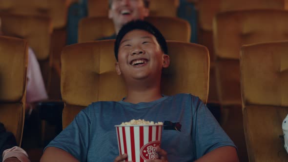Handsome cheerful young asian boy laughing while watching film in movie theater. alt