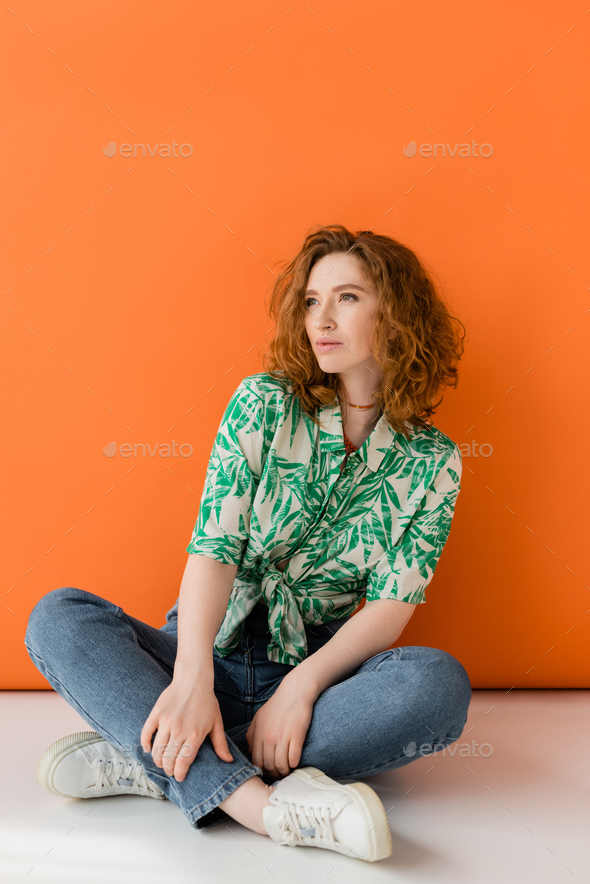 Confident young redhead woman in modern blouse with floral pattern and jeans looking away