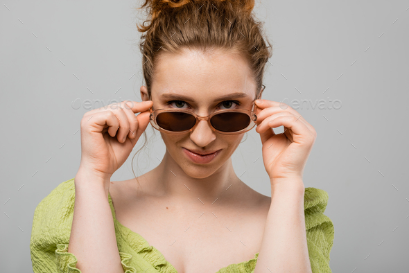 Stylish young redhead woman with natural makeup wearing green blouse and touching sunglasses