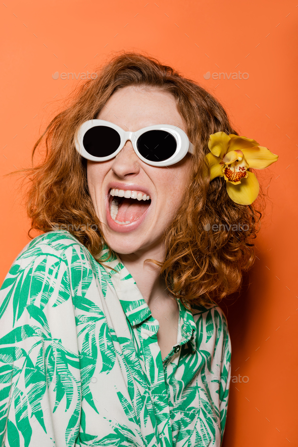 Excited redhead woman with natural makeup and orchid flower wearing sunglasses and blouse