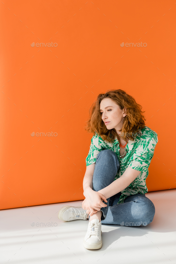 Full length of young red haired woman in modern blouse with floral pattern and jeans sitting