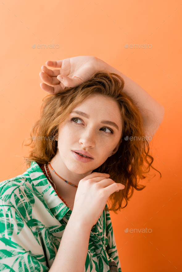 Portrait of dreamy and stylish redhead woman in modern blouse with floral pattern looking away and
