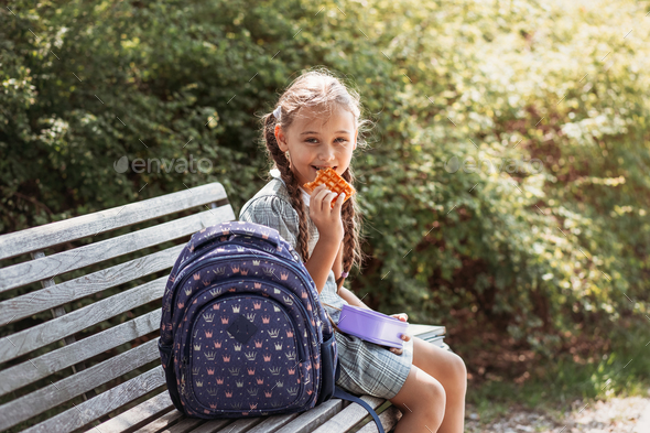  Cute little school girl sitting on bench in school yard and eating lunch outdoor.