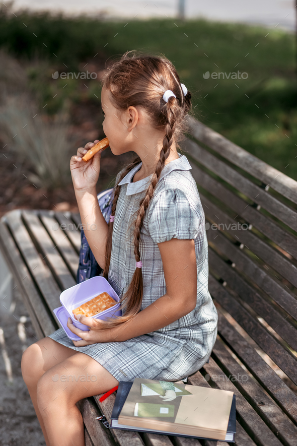 Cute little school girl sitting on bench in school yard and eating lunch outdoor.