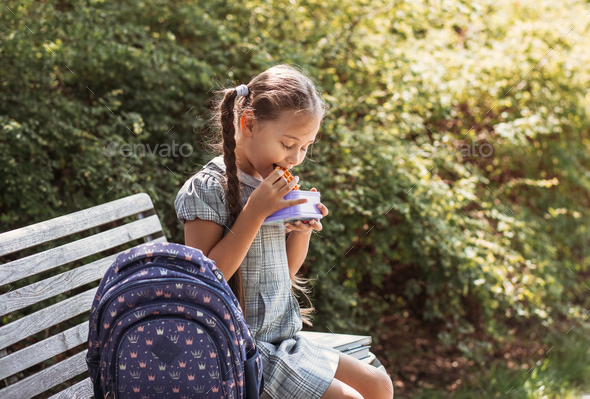 Cute little school girl sitting on bench in school yard and eating lunch outdoor.