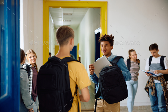 Happy black student taking laptop out of his backpack while talking to classmate at high school.