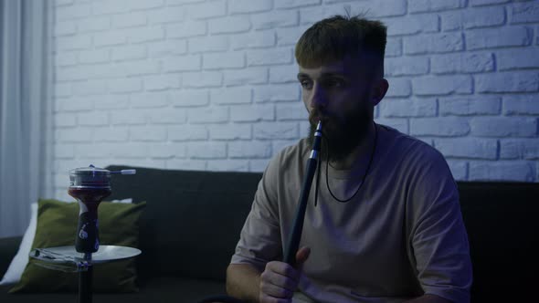 A Man is Sitting on the Couch at Home and Smoking a Hookah alt