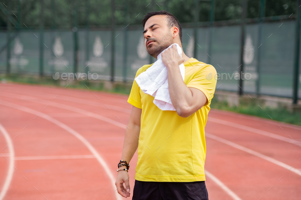 Exhausted sportsman wiping sweaty neck with towel standing on running track after jogging