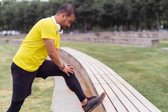 urban park, flexible man in black leggings leans on bench, doing exercises  to stretch leg muscles Stock Photo by demopicture