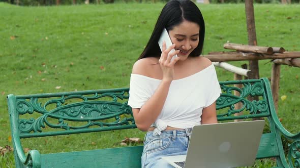 Young Asian woman using a laptop and using smartphone at a public park.