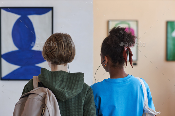Back view of two teens looking at art in art gallery sharing earphones