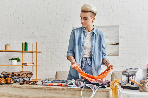 tattooed and casually styled woman sorting wardrobe items near electric toaster and cezve in modern