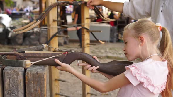 Blond Child Girl Aims and Shoots with Crossbow at Open Air Amusement Park Medium Shot alt