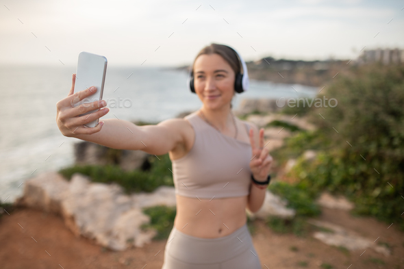 Happy millennial caucasian lady athlete in sportswear, wireless headphones makes peace sign, selfie