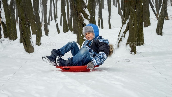 happy boy sliding down snow hill on sled outdoors in winter