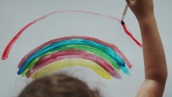 Little Girl Holds a Brush in Her Hand and Paints a Rainbow with Colored Paints on White Paper alt