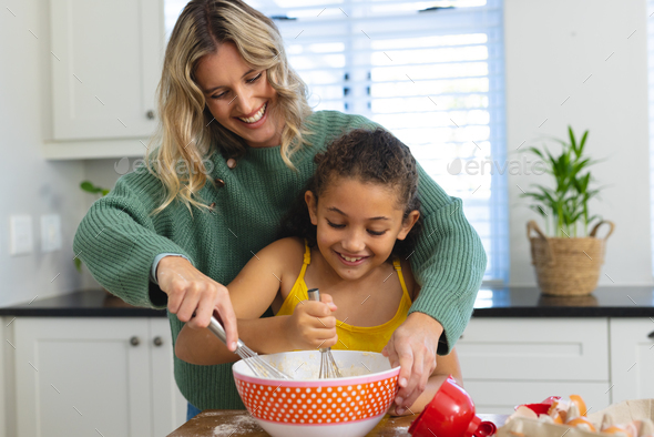 Cheerful multiracial mother teaching cute daughter to mix batter with wire whisk in bowl in kitchen